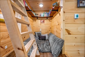 Une pièce chaleureuse, aménagée dans une cabane en bois, avec une échelle, une bibliothèque et un lit recouvert d’une couverture.