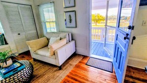 Una sala de estar con un sofá blanco, piso de madera, una puerta que da al exterior y una ventana con persianas.