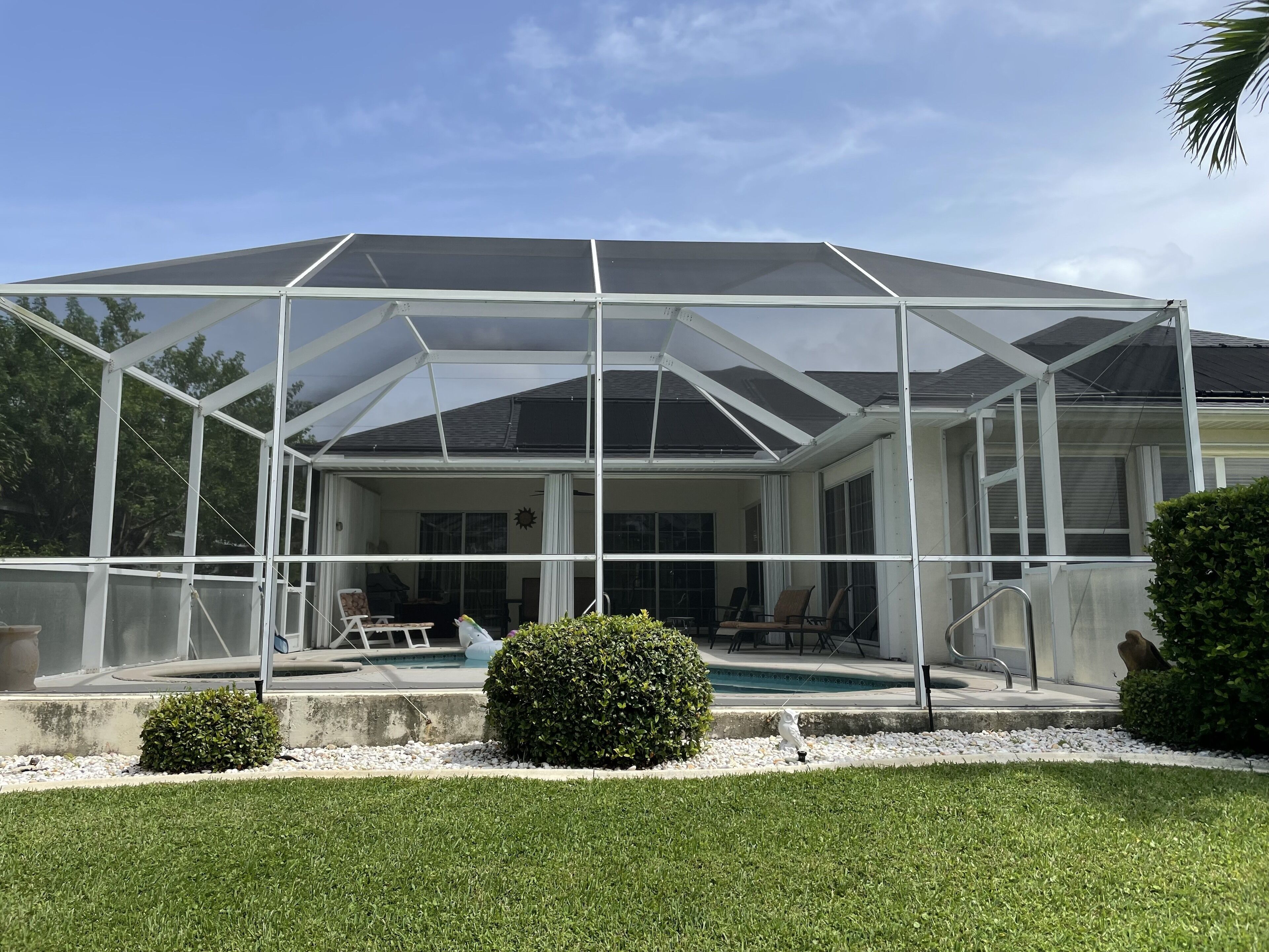A covered patio area with a pool, surrounded by a white fence and small bushes.