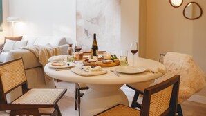 Een eetruimte met een ronde tafel gedekt voor vier personen, omringd door rieten stoelen. Op de tafel staan een fles wijn, glazen en borden met eten. De kamer heeft een modern design, met een groot abstract schilderij aan de muur en een zachte, pluizige fauteuil in de hoek.