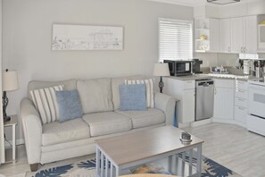 A modern living room with a sofa, a coffee table, and a kitchen area with white cabinets and appliances.