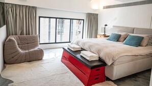 Une chambre moderne avec un grand lit, un banc de rangement rouge, un fauteuil confortable et une vue sur la ville grâce à une grande fenêtre.