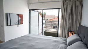 Une chambre avec un lit, une porte-fenêtre coulissante donnant sur un balcon, un téléviseur à écran plat et une vue sur les immeubles.