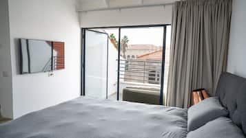 Une chambre avec un lit, une porte-fenêtre coulissante donnant sur un balcon, un téléviseur à écran plat et une vue sur les immeubles.