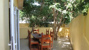 A patio with a table and chairs, surrounded by a yellow wall and trees. - Nice & private in our backyard with own entry & parking lot. (Willemstad)