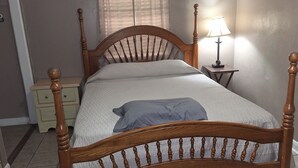 A bedroom with a wooden bed, a bedside table with a lamp, a nightstand, a window with curtains, and two framed pictures on the wall.