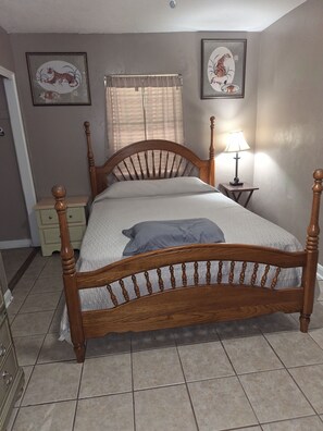 Un dormitorio con una cama de madera, una mesita de noche con lámpara, un tablero auxiliar, una ventana con cortinas y dos cuadros en la pared.