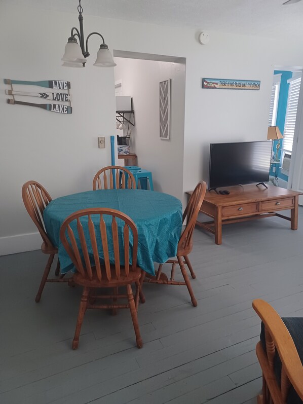 A dining area with a table covered in a teal tablecloth, surrounded by wooden chairs. A television is placed on a wooden cabinet to the right.