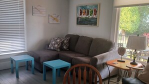 Ein Wohnzimmer mit Deckenventilator, einer Couch, einem Couchtisch, einem Sessel und einem Fenster mit Jalousien.