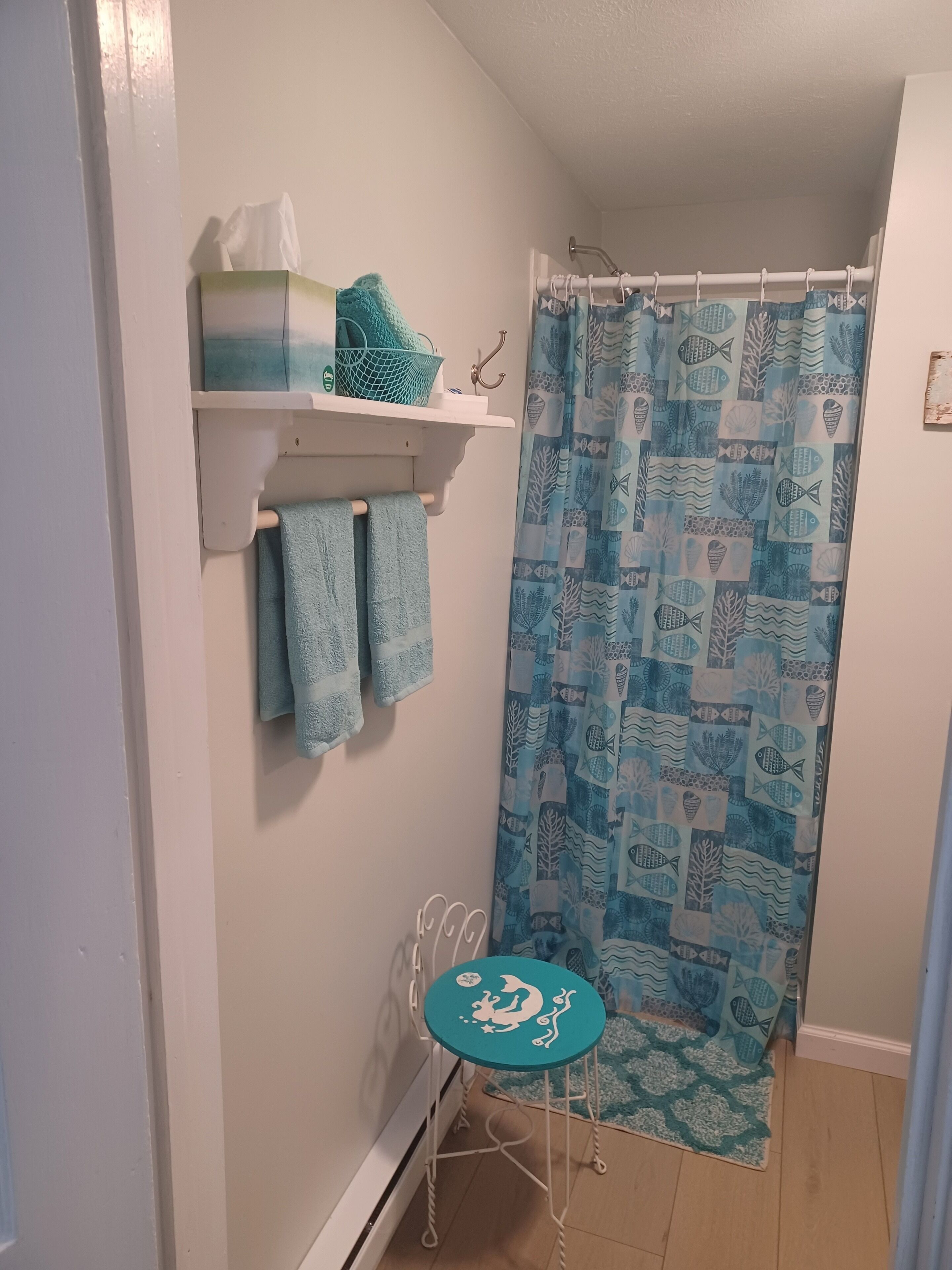 A bathroom with a shower curtain featuring a fish and coral pattern, a towel rack with two towels, a shelf with toiletries, and a small stool with a fish design.