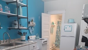 Eine kleine Küche mit weißen Schränken, einem Herd, einer Spüle und einem Kühlschrank. Eine blaue Wand und eine Tür zu einem anderen Raum.