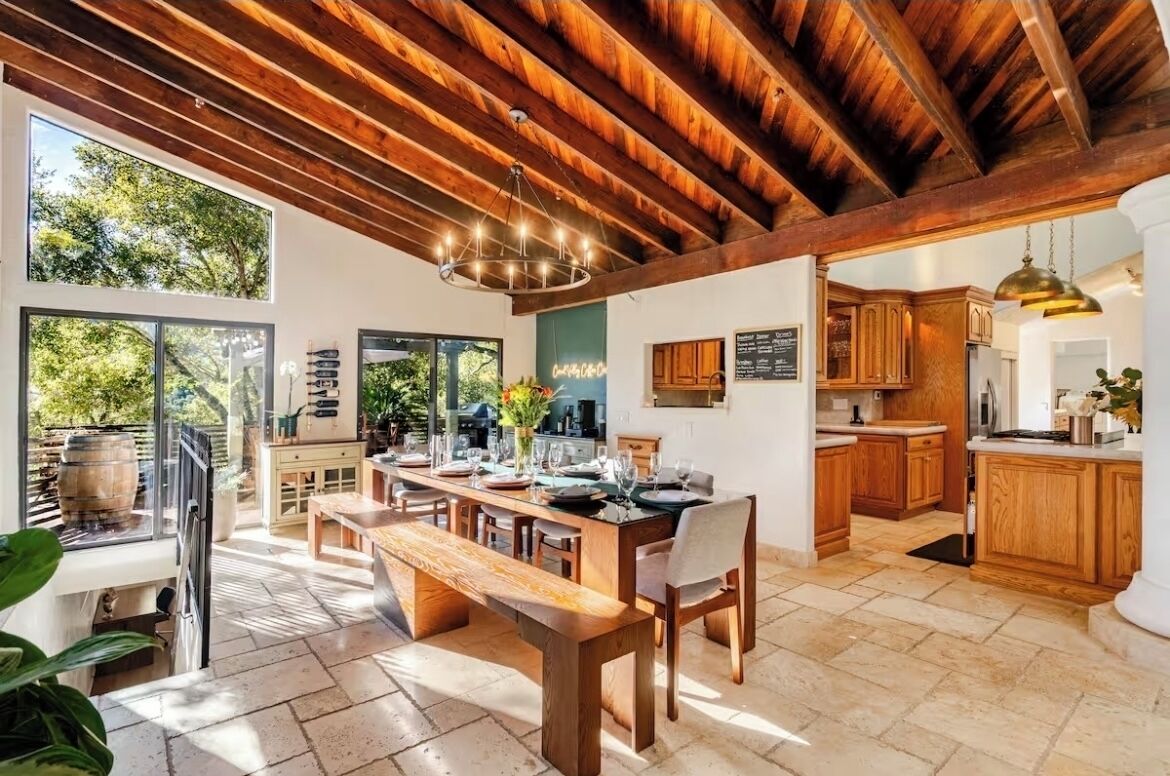 A spacious kitchen with wooden cabinets, a dining area with a bench, and large windows offering views of greenery.