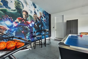 A room with a large superhero-themed mural, a ping pong table, and a basketball hoop.