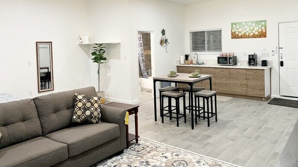 Ein modernes Wohnzimmer mit einer Couch, einem Couchtisch und einem Essbereich mit Tisch und Stühlen. Es gibt eine Küchenzeile mit einer Mikrowelle und einem Kühlschrank.