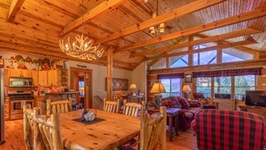 Interior rústico de una cabaña con una mesa de comedor de madera, sillas, chimenea y una zona de estar acogedora con sofá y televisión.