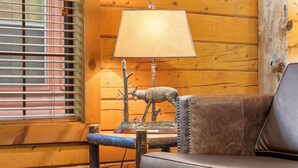 A wooden cabin interior with a leather sofa, a lamp with a deer sculpture, and a window with blinds. - Cozy Cabin for Two near Shows! Whirlpool Tub, Gas Fireplace, Private Balcony (Branson)