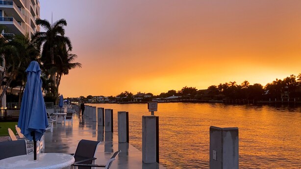 Een terras aan het water met tafels en stoelen, een uitzicht op de ondergaande zon over het water, en een rij palmbomen.