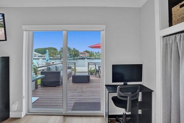 A room with a sliding glass door leading to a deck with outdoor furniture, a TV, and a desk. - Enjoy scenic sunsets on your private deck. Modern condo minutes to the beach. (Fort Lauderdale)