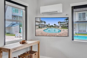 A room with a large TV displaying a pool view, a wooden table, and an air conditioning unit.
