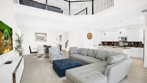 A spacious living area with a large grey sectional sofa, a blue ottoman, and a dining area with a white table and chairs. A kitchen with white cabinets and modern appliances is visible in the background. - Championsgate Resort 5 Bedroom Private Pool Home (Davenport)
