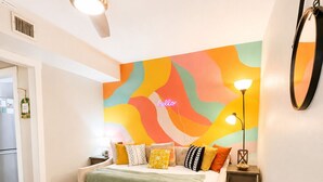 A living room with a sofa, a coffee table, a ceiling fan, a colorful abstract wall, and a small side table with a lamp. - Central 1BDR+PetsOK+Extended Stay Discounts (Corpus Christi)