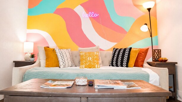 A room with a colorful abstract wall art, a sofa with patterned cushions, a wooden coffee table with magazines, and a neon sign that reads 'hello'. - Central 1BDR+PetsOK+Extended Stay Discounts (Corpus Christi)