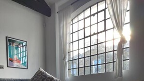 Ein Zimmer mit einer Couch, einem Sessel, einem Glastisch mit einem Buch, einem Fenster mit Vorhängen und einem gerahmten Bild an der Wand.