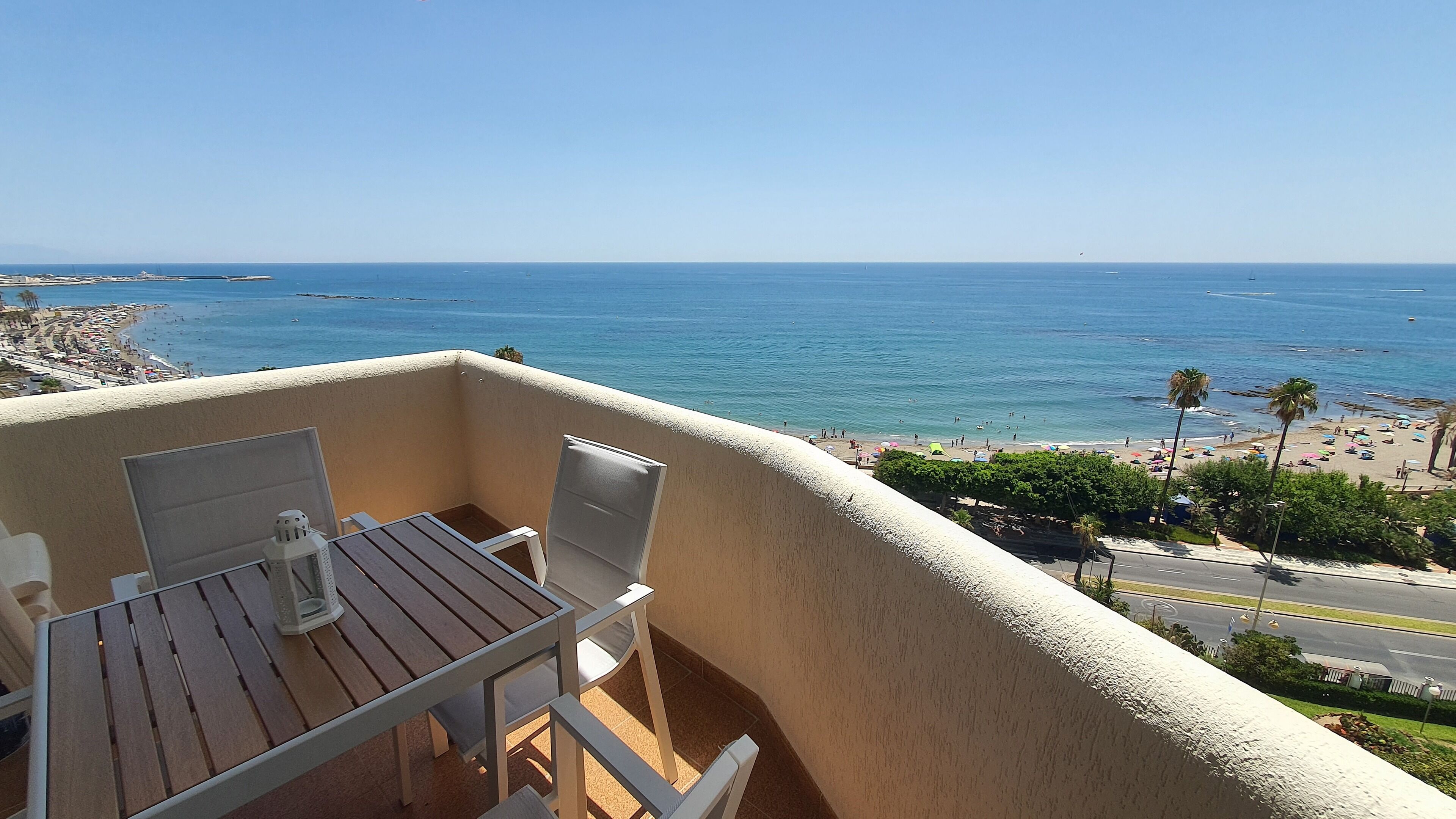A balcony with a wooden table and chairs, overlooking a beach with palm trees and a clear blue sky.