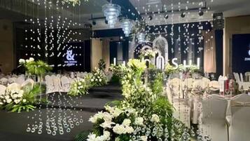 Un salón de banquetes con un escenario, arreglos florales blancos e iluminación colgante.