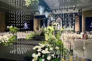 Un salón de banquetes con un escenario, arreglos florales blancos e iluminación colgante.