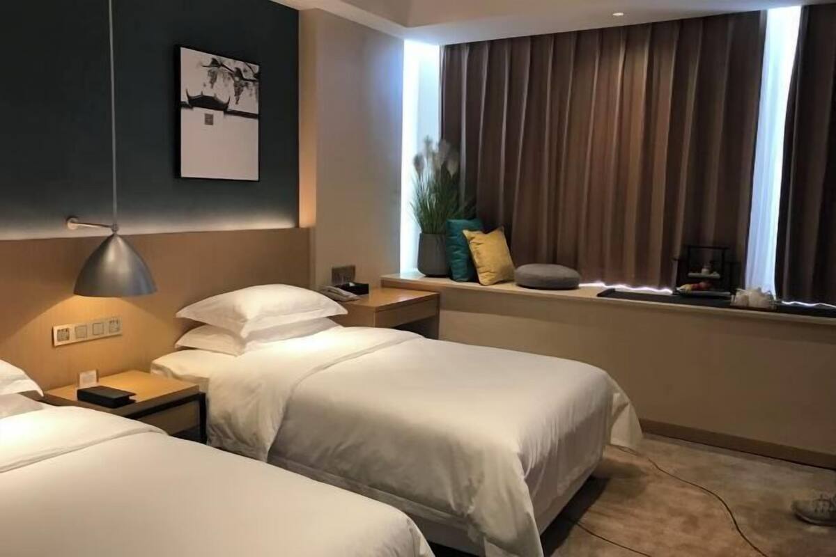 Quarto de hotel com duas camas, uma mesa de cabeceira com abajur, uma janela com cortinas e um quadro pendurado na parede.