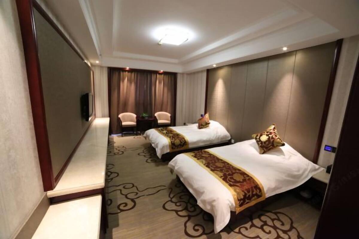 A hotel room with two beds, a sitting area with a chair, a TV, and a large window with curtains.