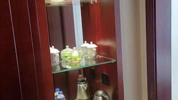 Habitación de hotel con repisa de vidrio que sostiene varios objetos, una tetera y una botella de agua.