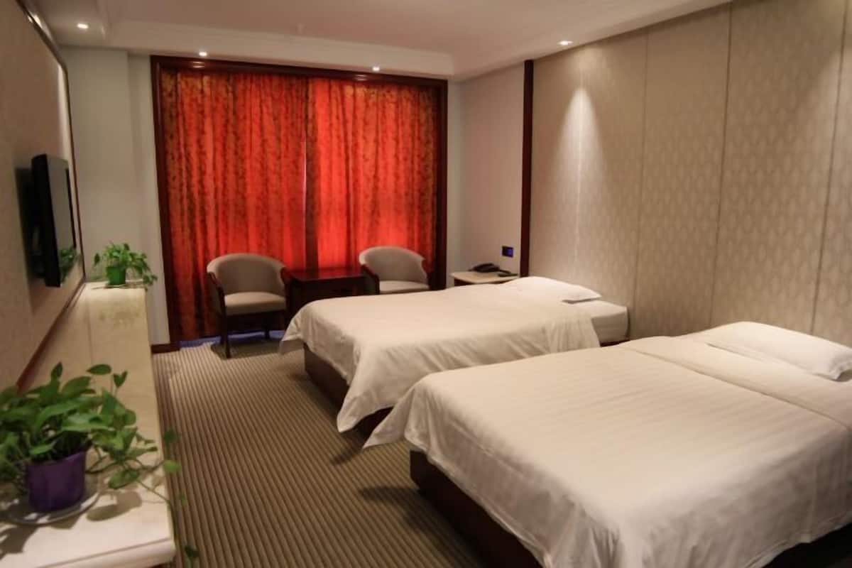 A hotel room with two beds, a desk, two chairs, a TV, and a large window with red curtains.