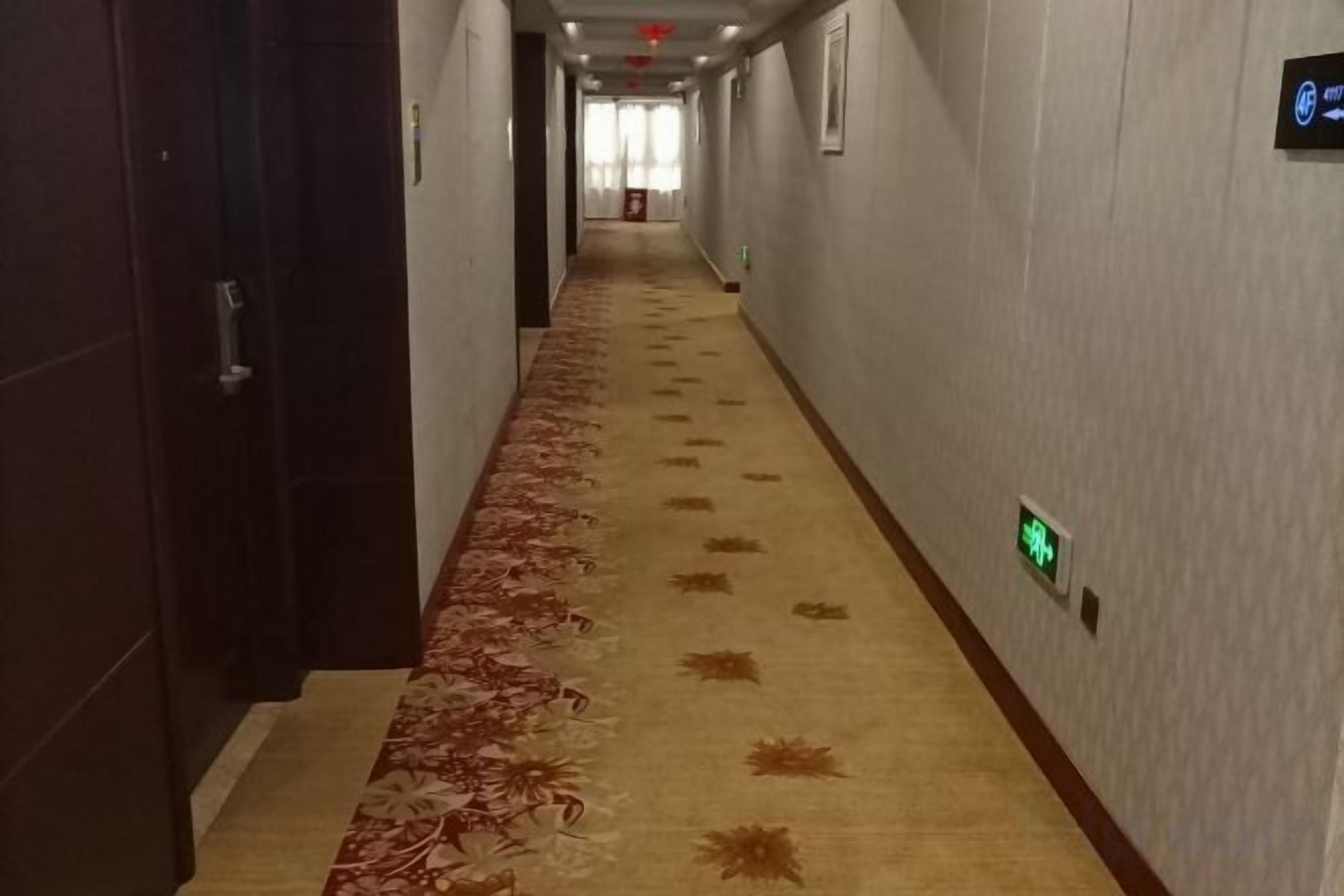 Un pasillo de hotel con suelo alfombrado, puertas a la izquierda y señalización de salida de emergencia a la derecha.