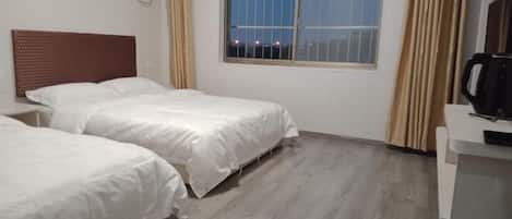 A hotel room with two single beds, a window with a view of the city at night, a wooden floor, and a small desk with a coffee maker.