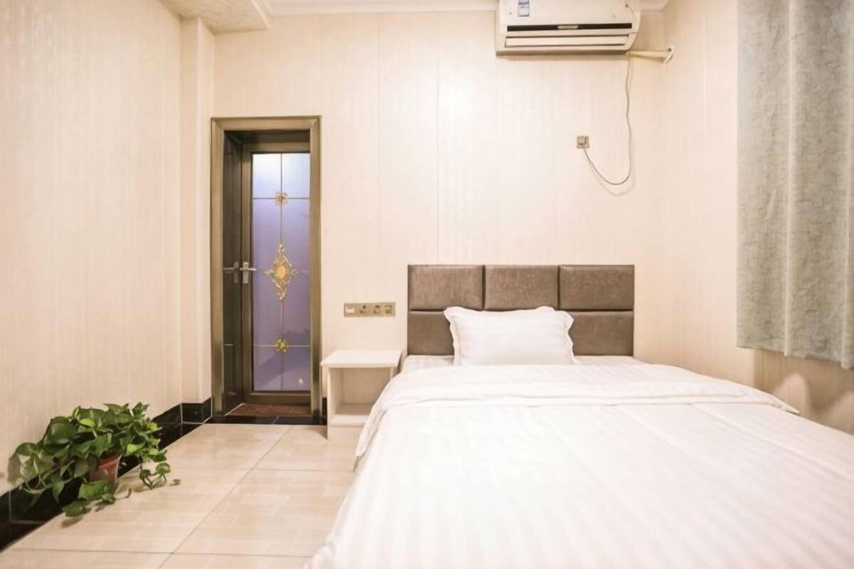 A hotel room with a bed, bedside table, potted plant, and an air conditioning unit.
