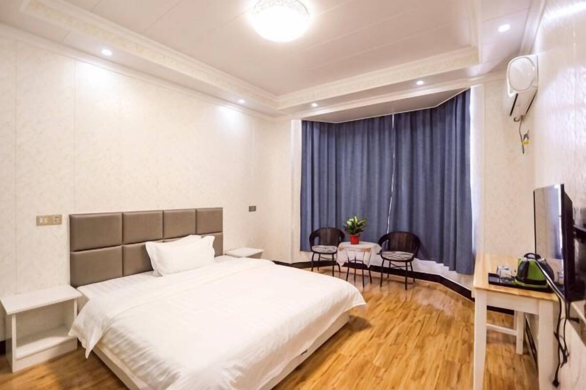 A hotel room with a bed, a desk, a chair, a small table, a plant, and a television.