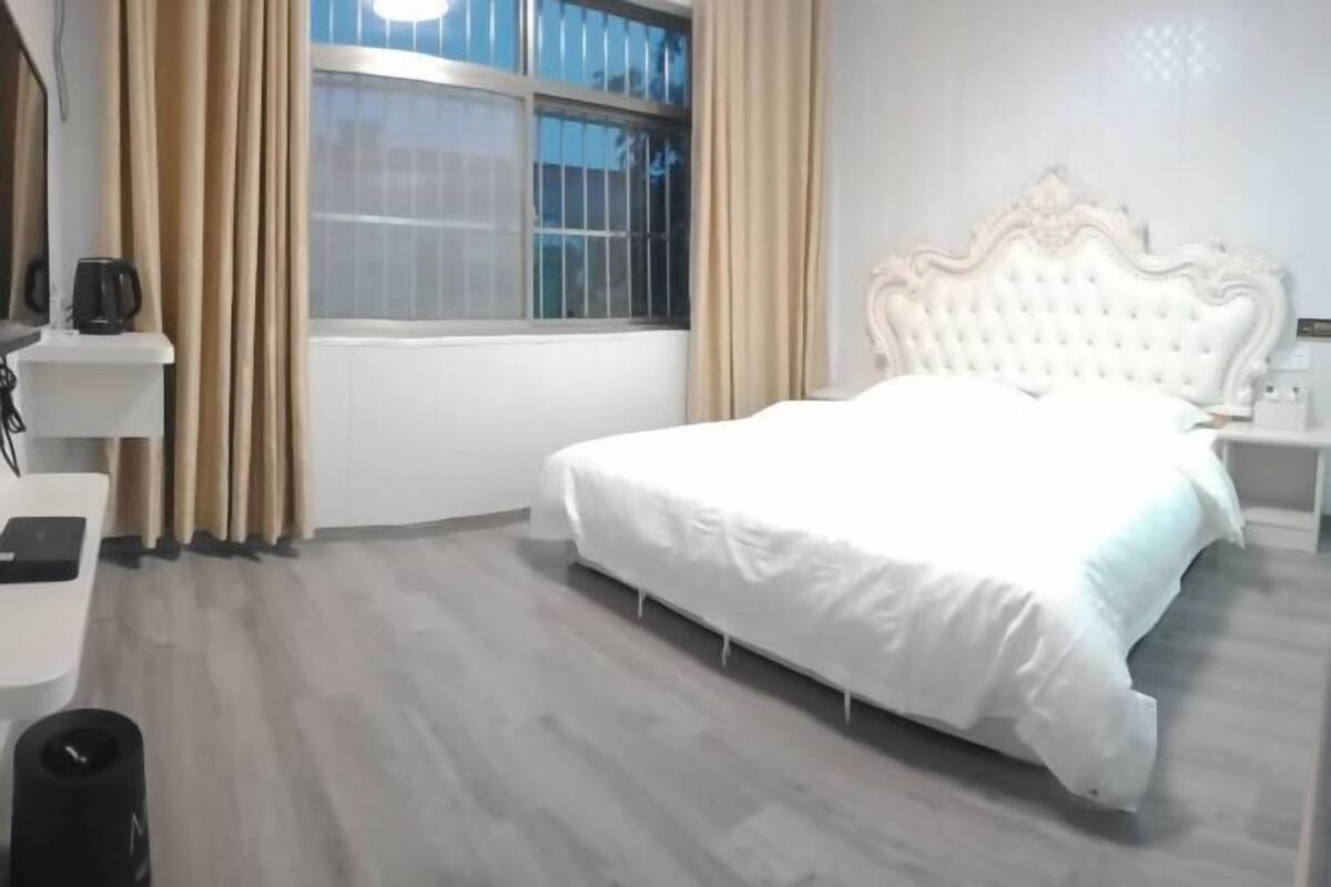 A bedroom with a large bed, a bedside table, a window with curtains, and a radiator.