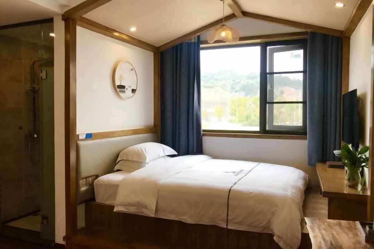 Un dormitorio con cama, una mesita de noche con una planta, una ventana con cortinas y un rincón de ducha.