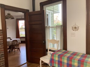 A room with a wooden door, a window with curtains, a small table with a colorful tablecloth, and a chair.