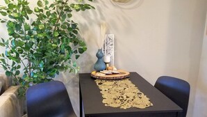A dining area with a black table, two black chairs, a gold table runner, a potted plant, and a wall art piece.
