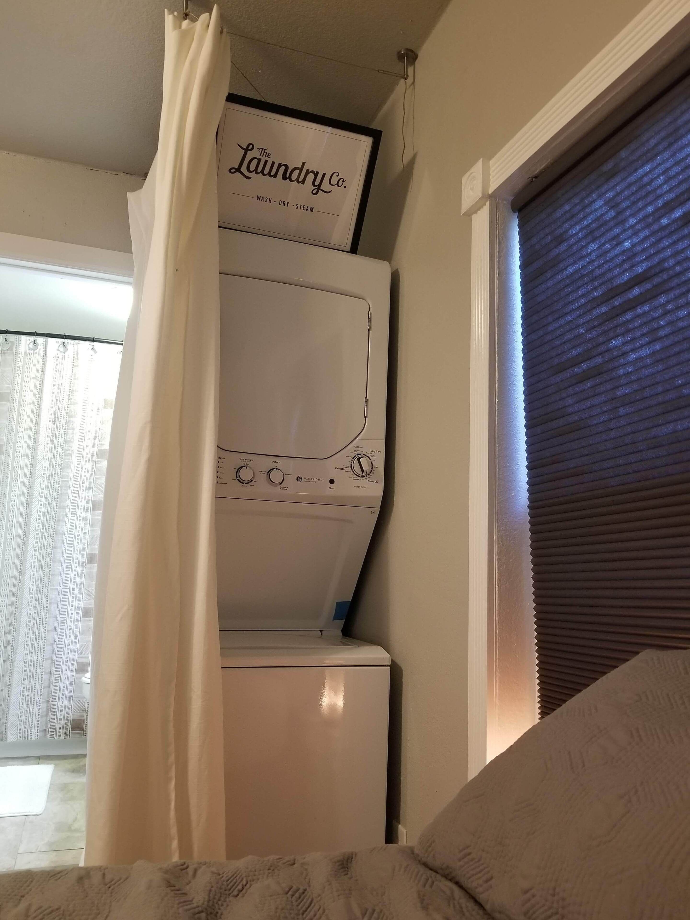 A laundry room with a white washing machine, a framed sign above it, and a window with blinds.