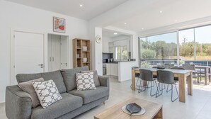 Una sala de estar moderna con un sofá gris, una mesa de centro de madera y un comedor con un amplio ventanal que ofrece vistas al exterior.
