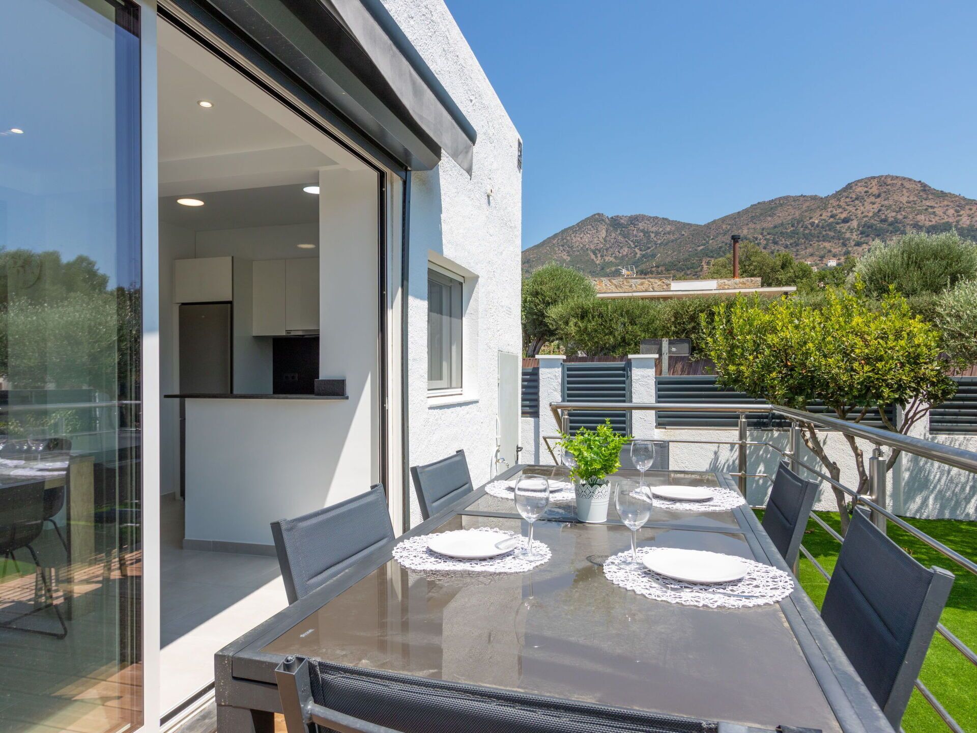A modern outdoor dining area with a glass table and chairs, overlooking a mountainous landscape.