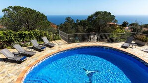 Una piscina con borde de azulejos, rodeada de sillas de descanso y una valla metálica, con vistas a un paisaje costero.
