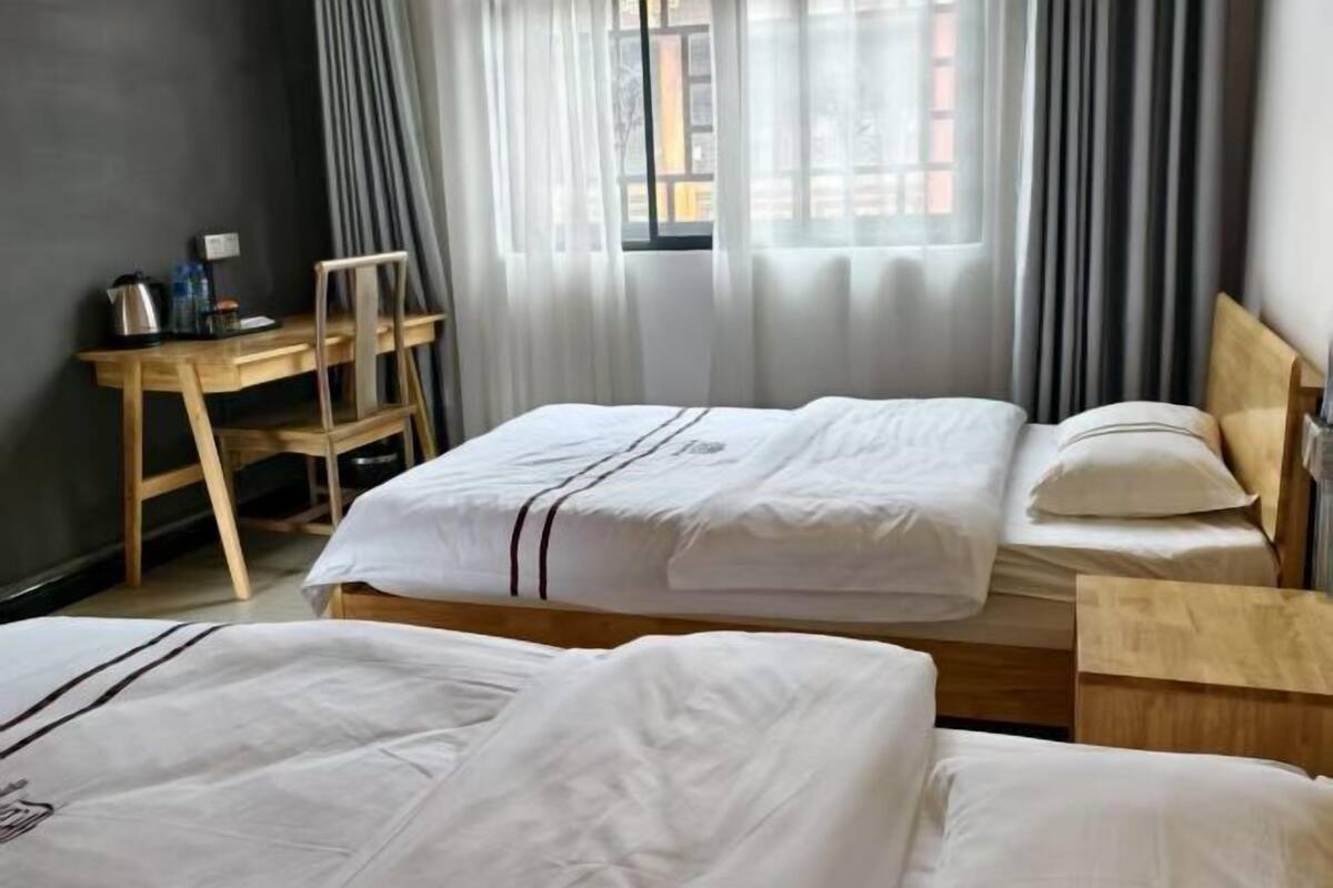 Habitación de hotel con dos camas, un escritorio de madera, una silla y un ventanal con cortinas.