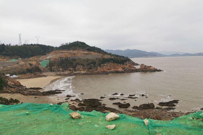 A coastal landscape with a beach, rocky shore, and a hill with sparse vegetation.