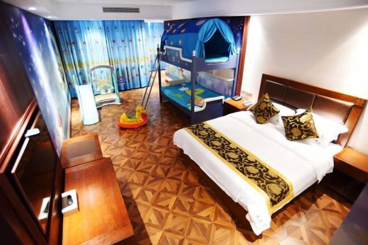 A hotel room with a bunk bed, a wooden desk, a small table, a nightstand, a bed with patterned bedding, and a colorful mural on the wall.
