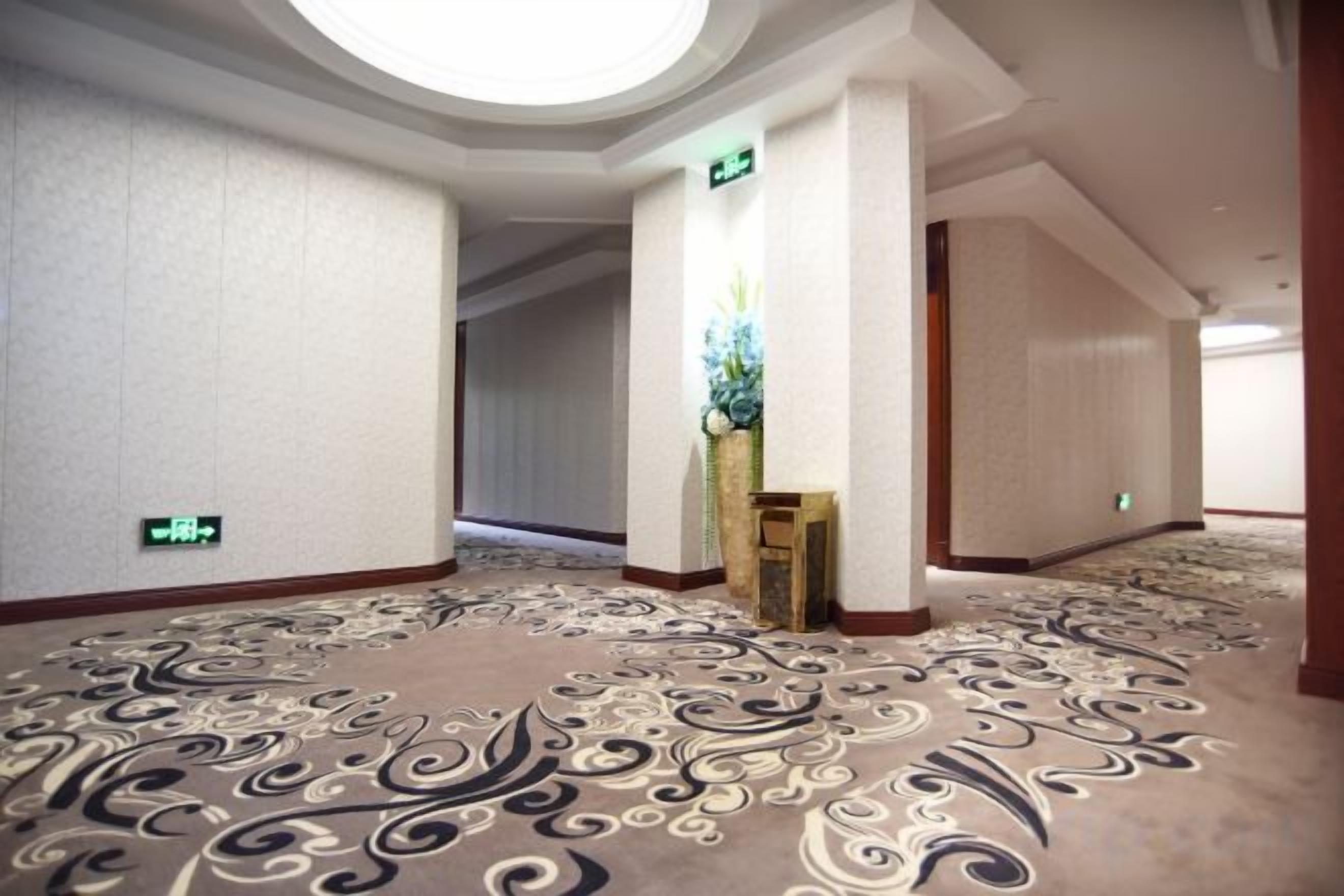 A hotel corridor with patterned carpet, a wooden reception desk, and a wall-mounted exit sign.
